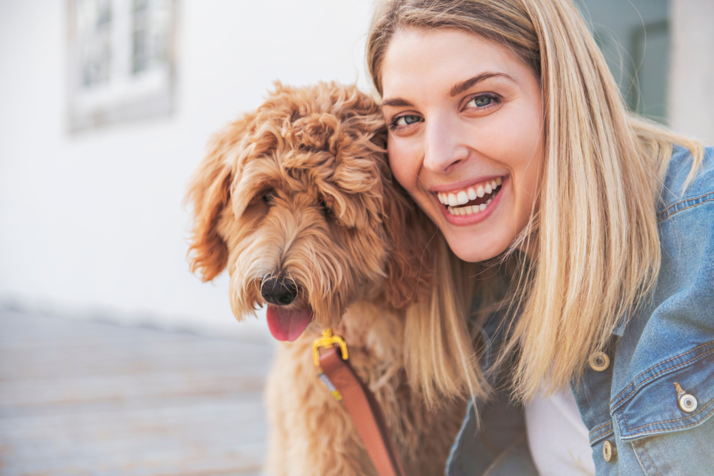 A smiling blonde woman hugs her Goldendoodle, happy that she found her furry friend. 