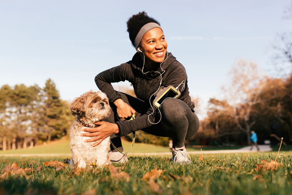 A happy, healthy black woman bends down during her daily jog to pet her Shih Tzu on a sunny autumn day.