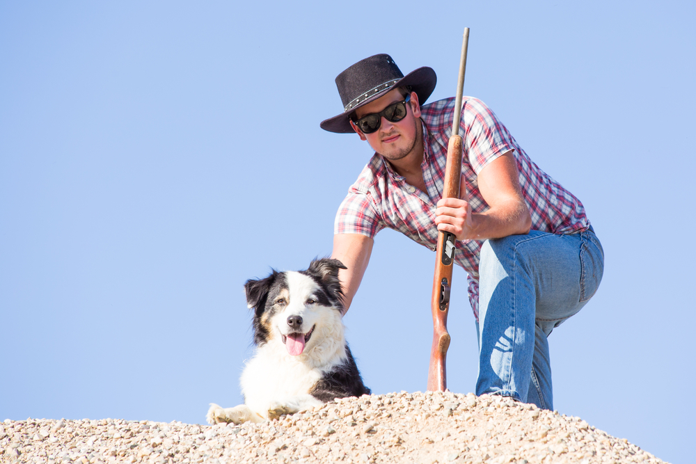 A cowboy rancher crouches with his rifle next to a purebred Australian Shepherd, as Australian Shepherd dogs were the original cowboy and rancher breed of choice in the American West.