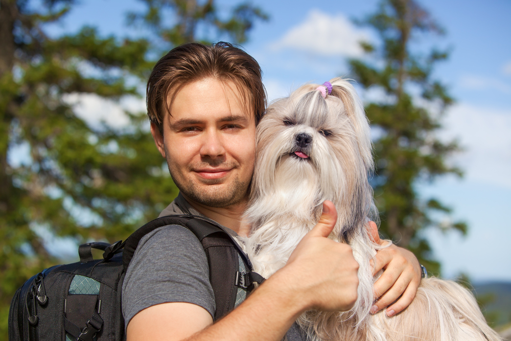 A long haired Shih Tzu smiles as its favorite person, a man, holds and hugs it while on a hike through the mountains.
